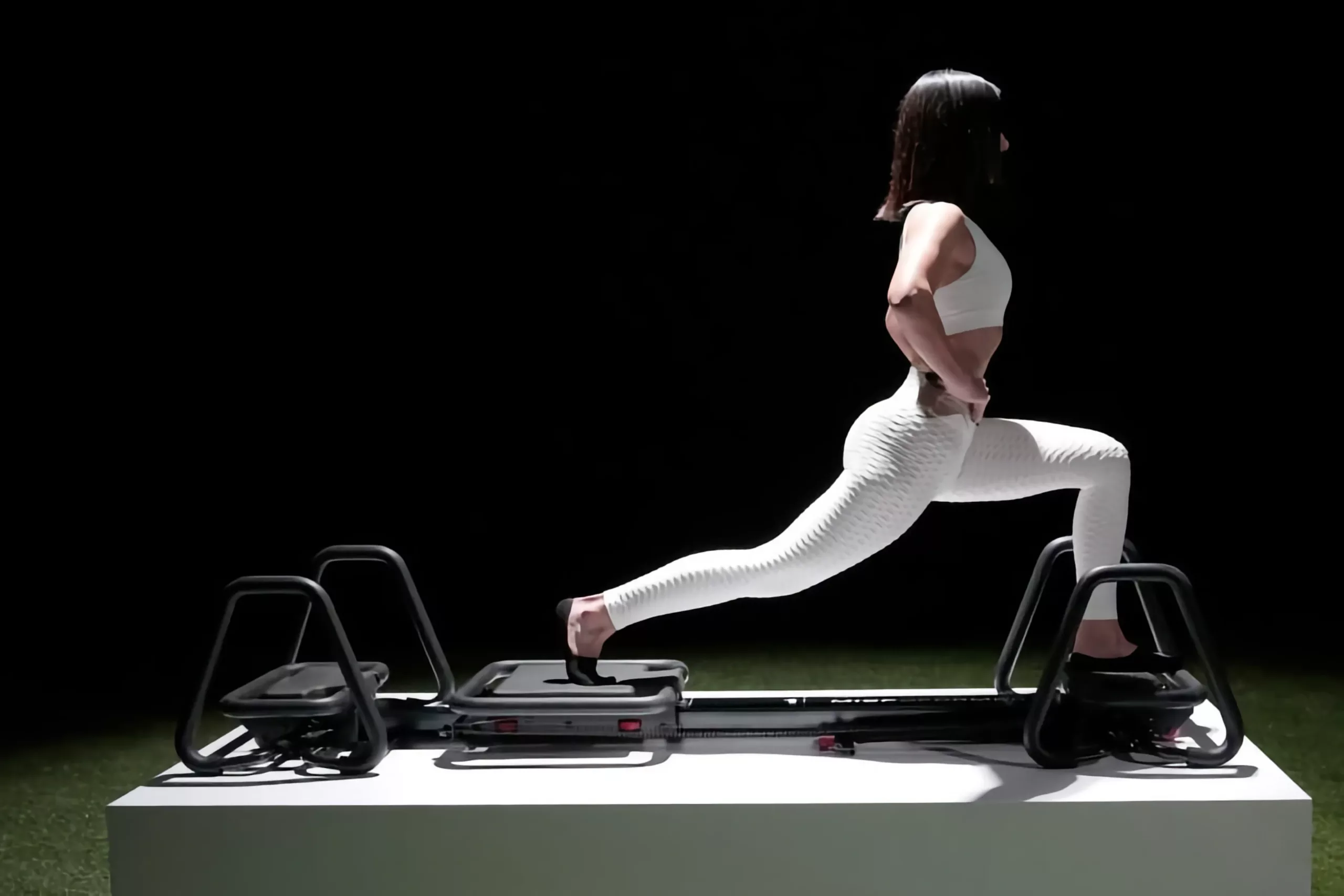 Femme athlétique en brassière et leggings blancs en fente arrière sur un appareil de fitness à glissière avec poignées, fond noir – renforcement des quadriceps, fessiers et équilibre dynamique.