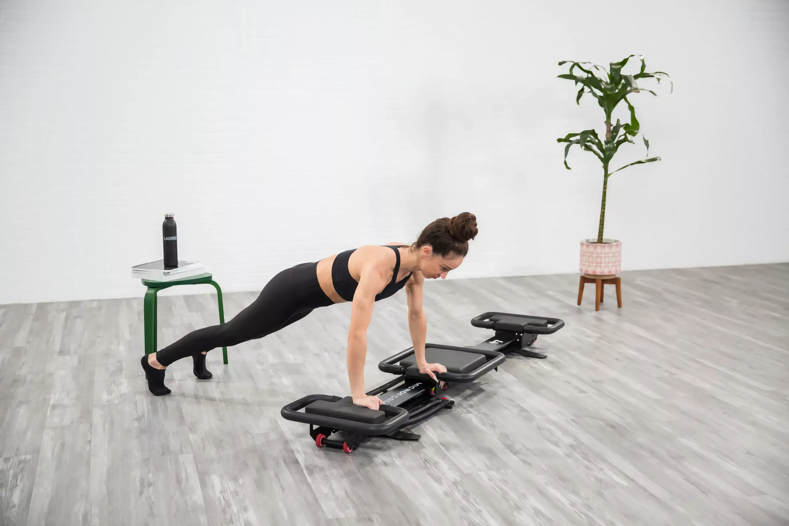 Femme en brassière et leggings noirs effectuant une planche push-up sur un appareil de fitness glissant portable à poignées, intérieur épuré avec sol parquet gris, mur blanc, plante en pot et tabouret vert avec gourde.