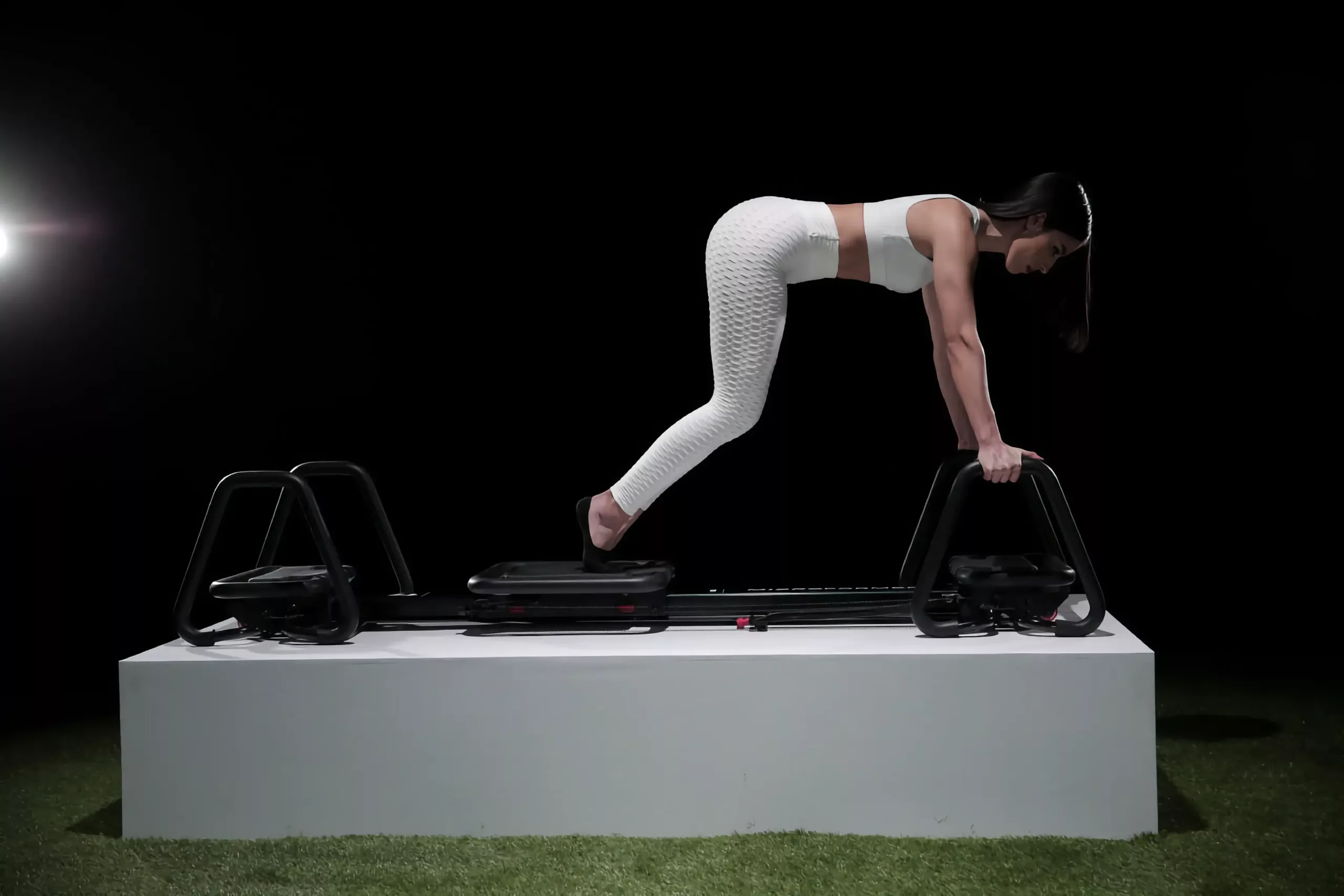 Femme athlétique en leggings et brassière blancs réalisant un exercice de planche dynamique sur un appareil de fitness à glissière avec poignées, fond noir – entraînement fonctionnel et gainage du tronc.