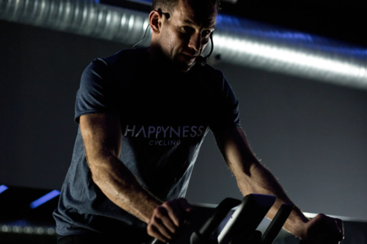 Coach de cycling en pleine séance, vêtu d’un t-shirt “Happyness Cycling” et équipé d’un micro-casque, concentré sur son vélo dans une salle à l’ambiance sombre avec éclairage d’ambiance.