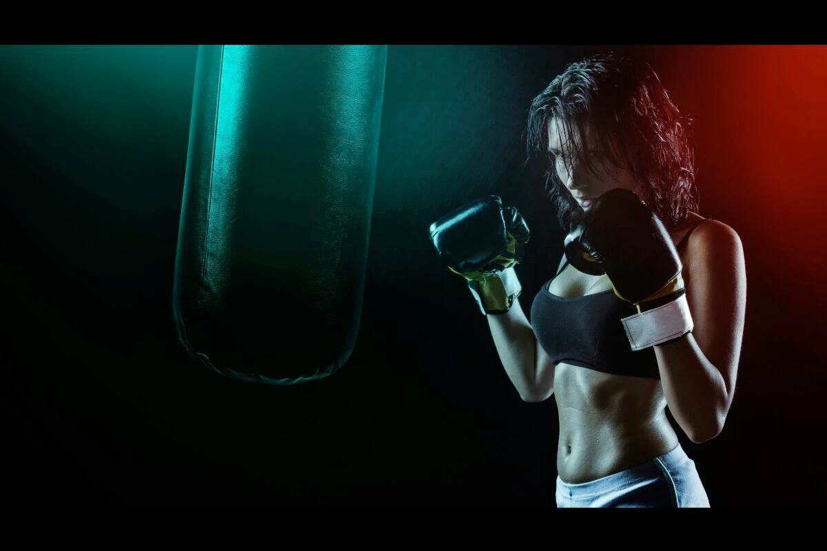Femme en tenue de boxe, concentrée face à un sac de frappe suspendu, dans une ambiance sombre rehaussée d’éclairages rouges et verts, symbolisant intensité et détermination.