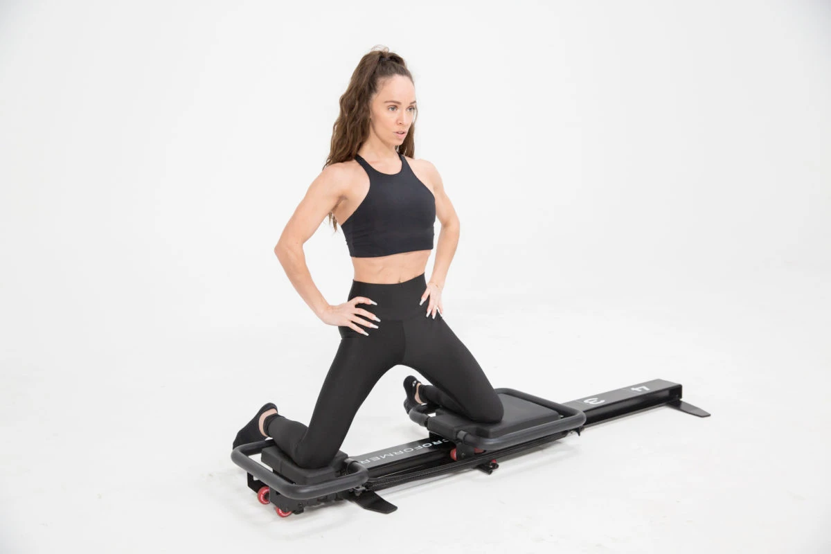 Femme en tenue de sport noire effectuant un exercice de renforcement musculaire sur une machine de type glute drive ou reformer, les genoux posés sur des plateformes coulissantes, sur fond blanc épuré.