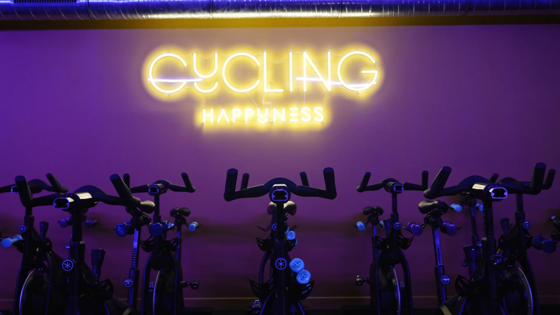 Salle de cycling avec plusieurs vélos de studio alignés sous une enseigne lumineuse jaune, dans une ambiance violette.