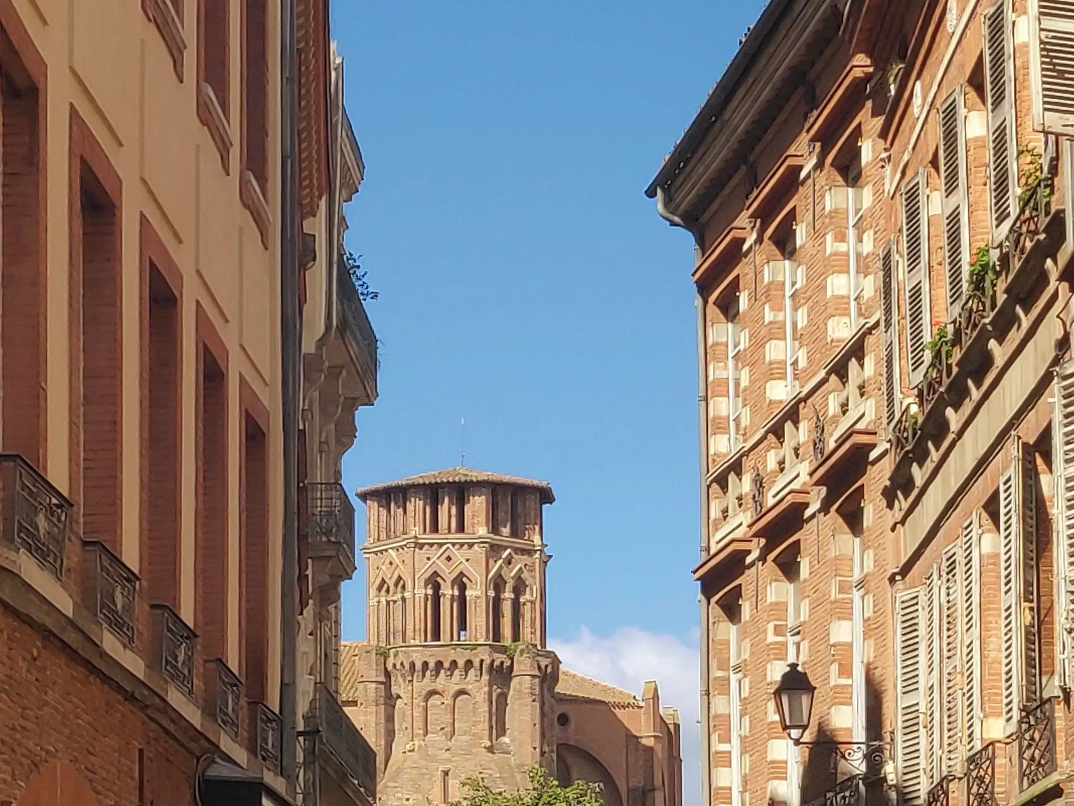 Rue étroite bordée d’immeubles en briques rouges, avec au centre une tour ancienne ornée de motifs gothiques sous un ciel bleu dégagé.